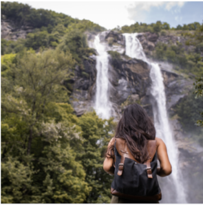 The back of a woman with a backpack facing a waterfall on a green cliffside